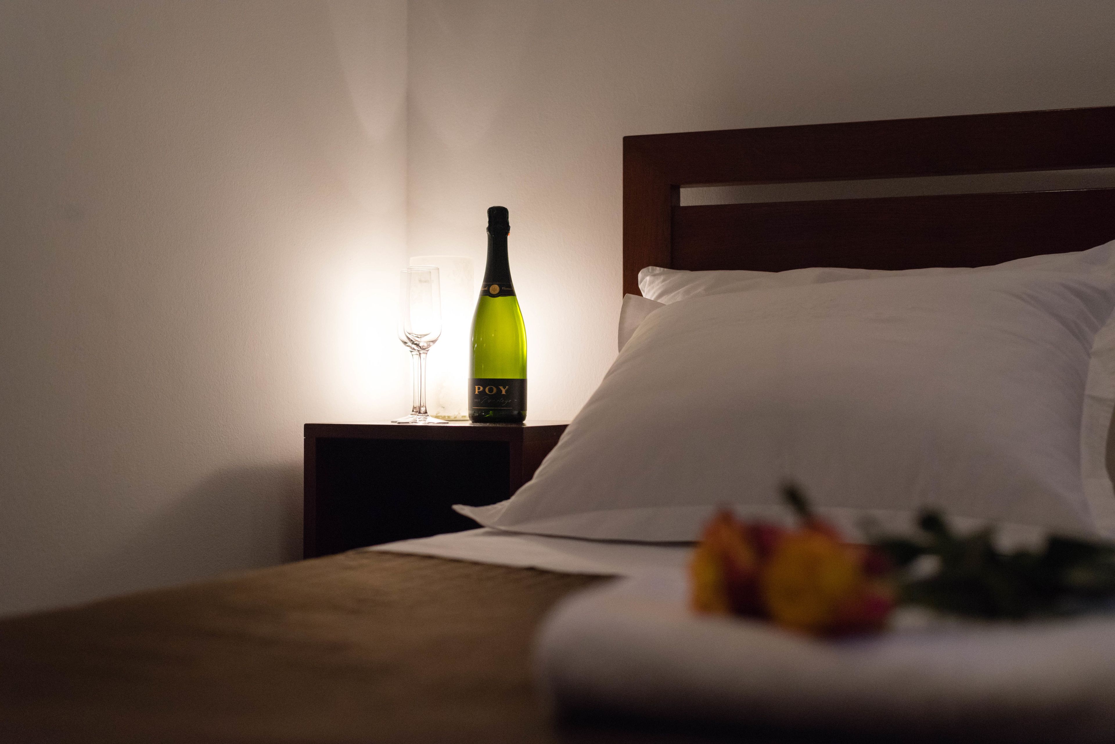 A hotel room with a neatly made bed, a bedside table with a bottle of wine and a glass, and a bouquet of flowers on the bed.