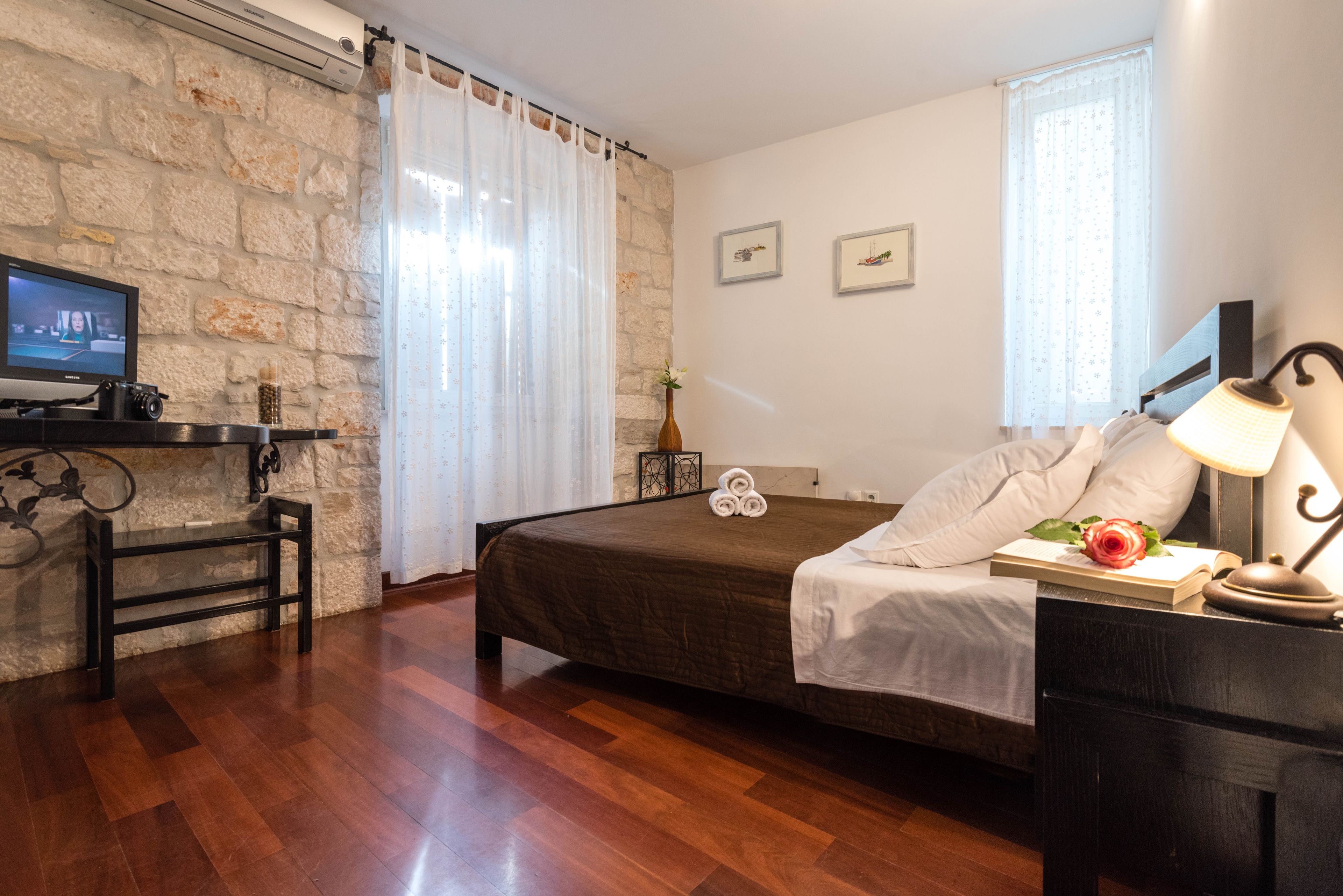 Un dormitorio con pared de piedra, suelo de madera, una cama con colcha marrón, una mesita de noche con lámpara, un televisor en un estante y una ventana con cortinas.