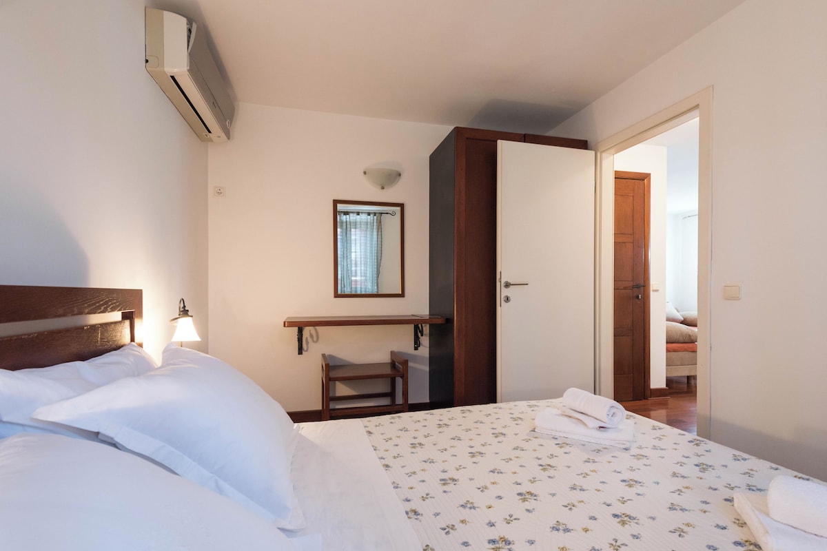 Habitación de hotel con cama, lámpara de noche, pequeño escritorio con silla, espejo y puerta que da a otra habitación.