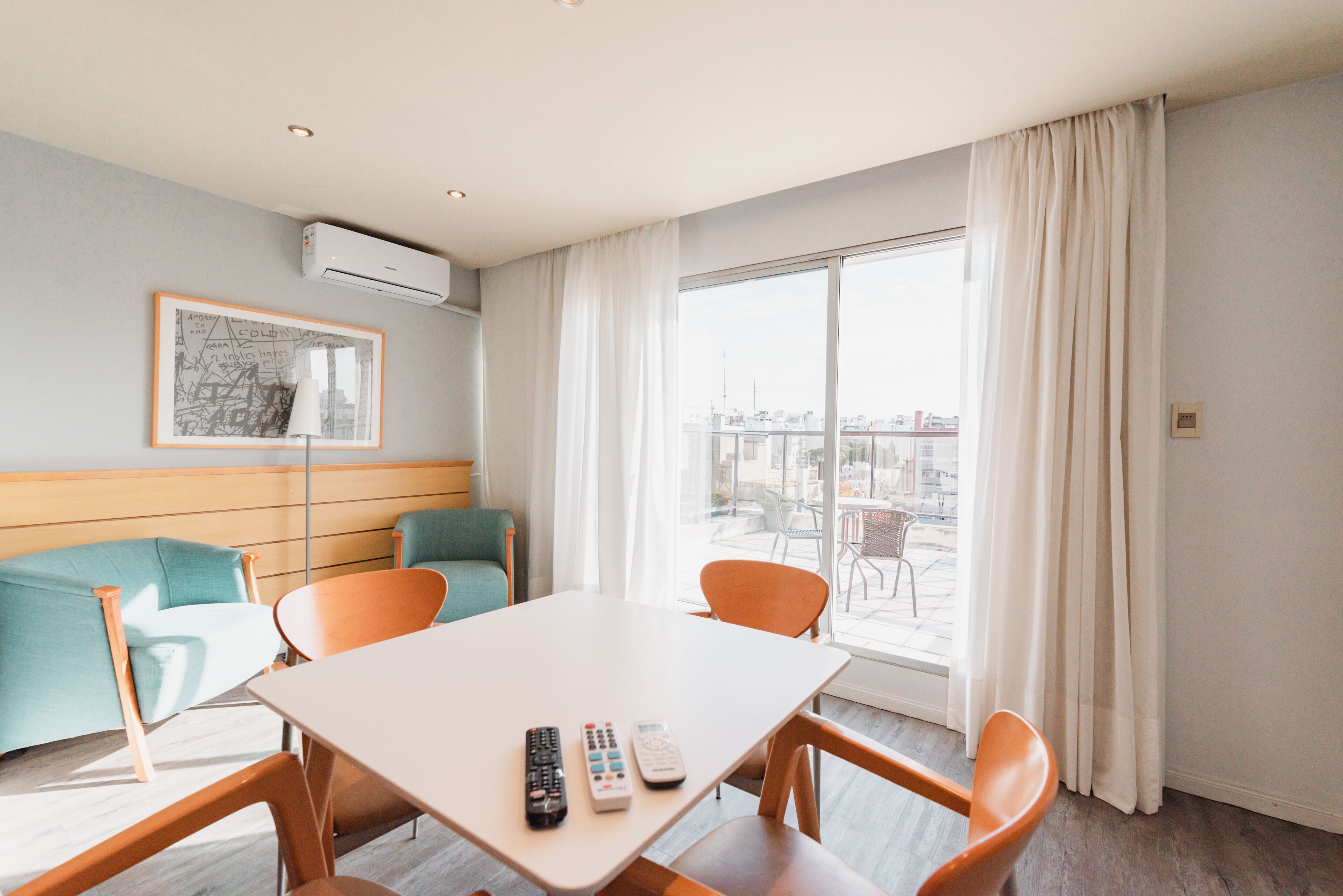 Habitación de hotel con mesa de comedor, sillas, sofá, un cuadro enmarcado y un ventanal con cortinas.