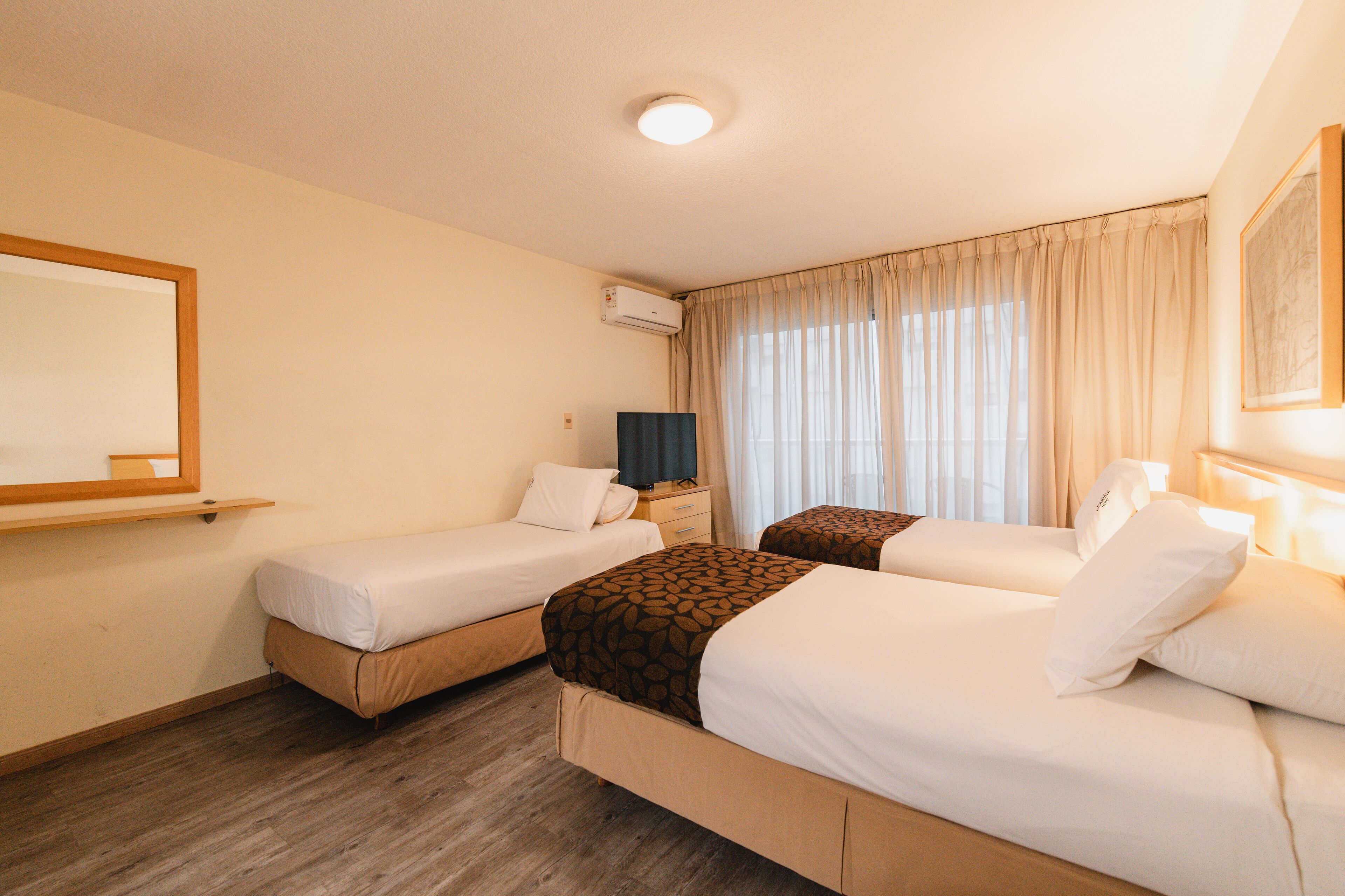 A hotel room with two single beds, a flat-screen TV, an air conditioning unit, and a large window with curtains.