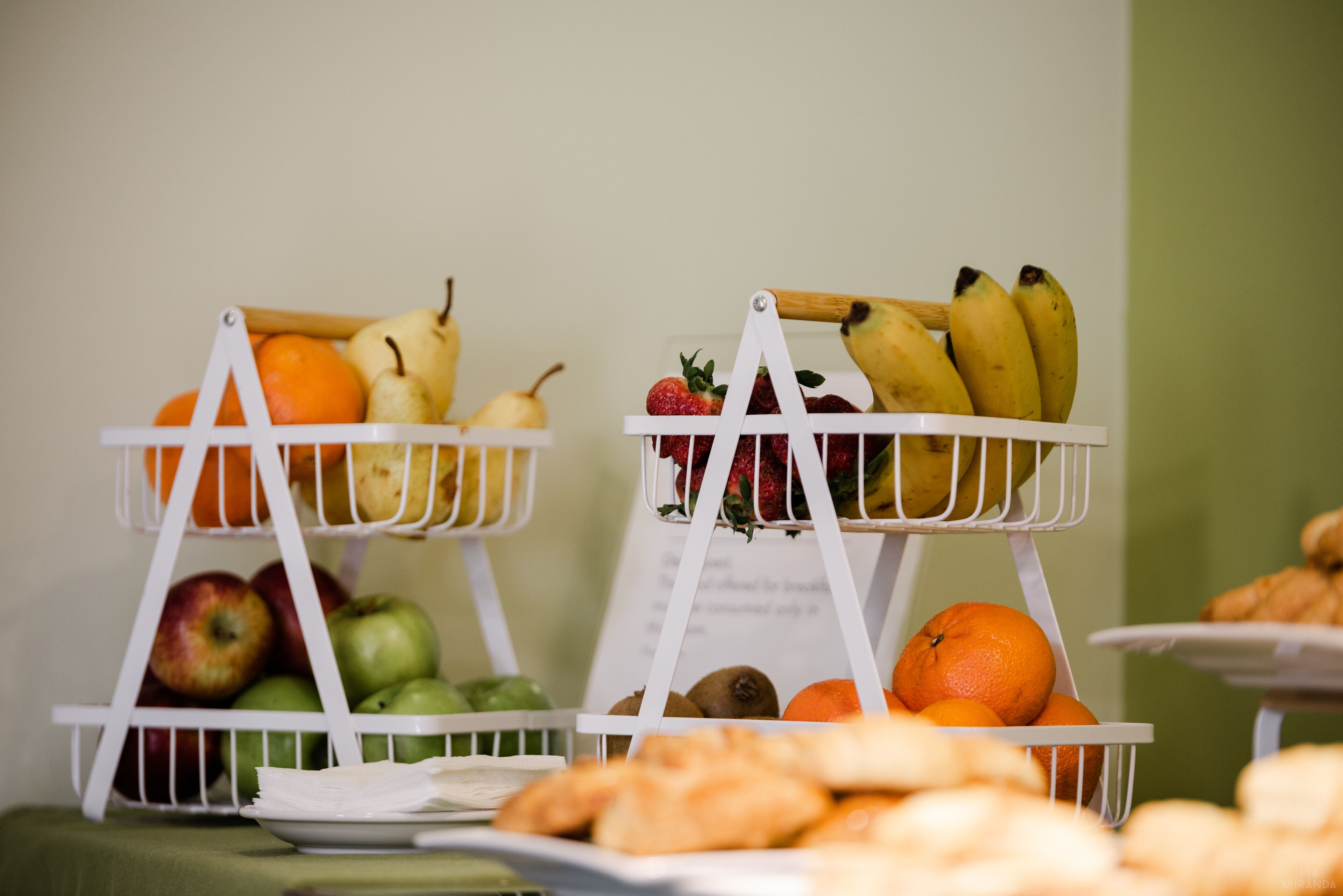 Dos cestas blancas de fruta sobre una mesa con diversas frutas y pasteles.