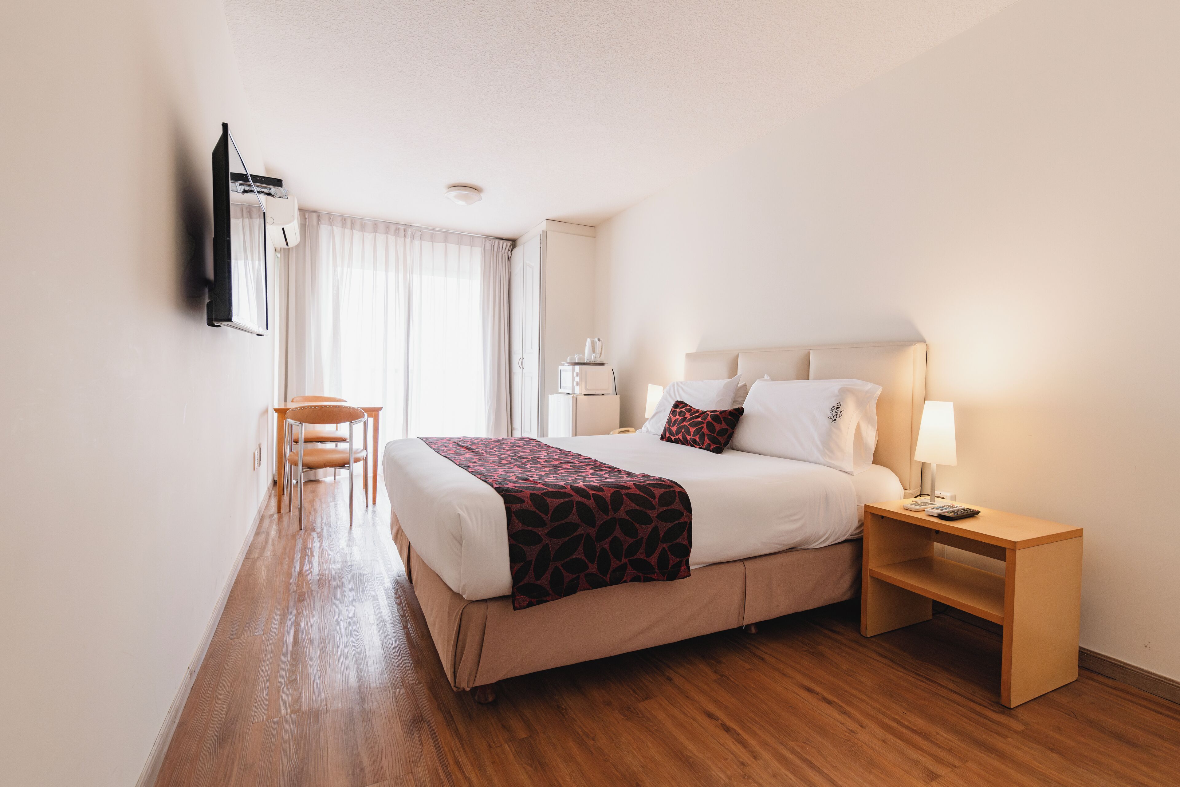 A hotel room with a bed, bedside table, lamp, and a TV mounted on the wall.