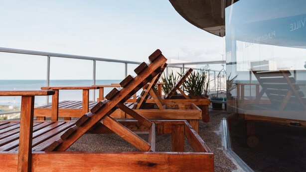 Outdoor wooden lounge chairs and tables on a terrace with a view of the ocean. - Punta Trouville Hotel (Montevideo)