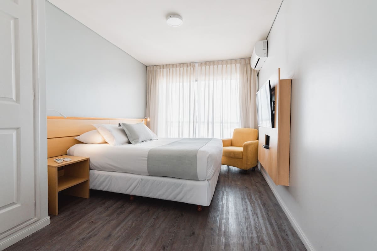 Una habitación de hotel moderna con una cama grande, una mesita de noche, una silla y un televisor montado.