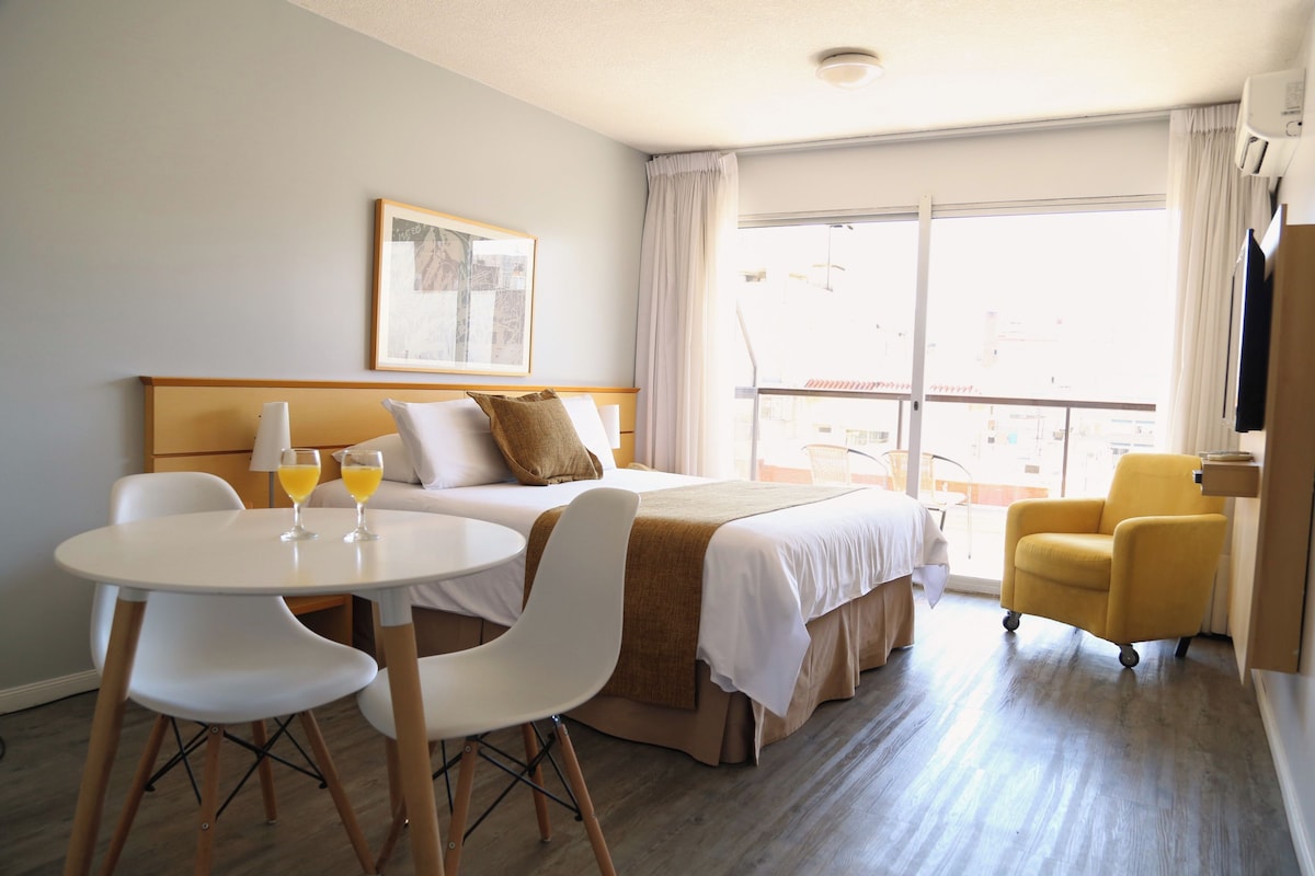 Une chambre d’hôtel avec un lit, une table ronde blanche et deux chaises, un fauteuil jaune et une grande fenêtre avec des rideaux.