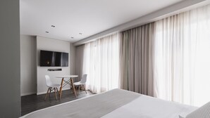 A modern hotel room with a large bed, a flat-screen TV mounted on the wall, a dining area with a table and chairs, and large windows with sheer curtains. - Punta Trouville Hotel (Montevideo)