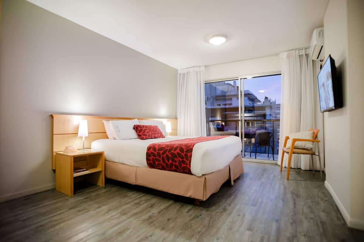 Habitación de hotel con cama, mesita de noche, silla y balcón con vistas a edificios.