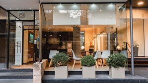 A hotel entrance with a glass facade, outdoor seating area with chairs and potted plants, and a staircase leading up to the entrance. - Punta Trouville Hotel (Montevideo)