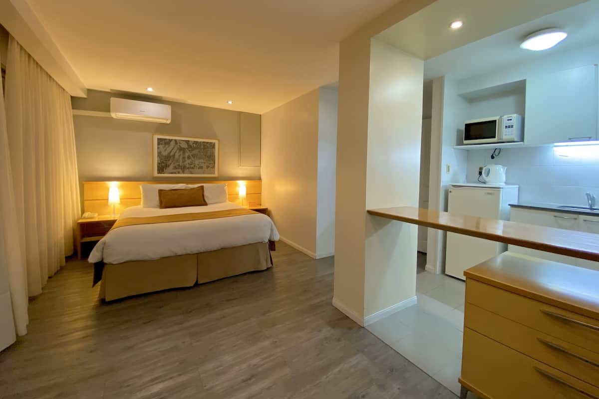 Habitación de hotel con cama, lámparas de noche, mesita auxiliar pequeña, microondas, hervidor de agua y cómoda de madera.