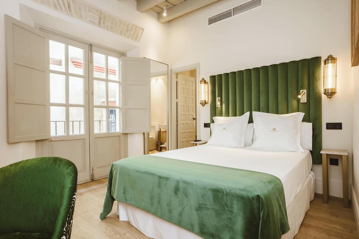A hotel room with a large bed, a green upholstered chair, a green headboard, and a nightstand with a lamp.