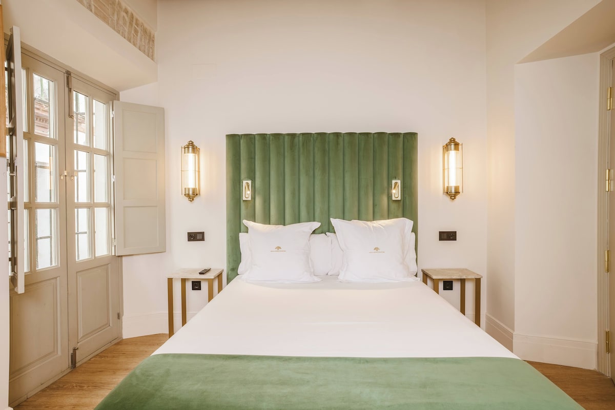 Habitación de hotel con una cama grande, cabecera verde, ropa de cama blanca, dos mesitas de noche y lámparas fijadas a la pared.