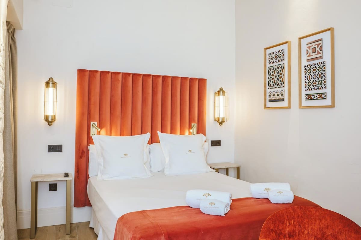 A hotel room with a large bed, two bedside tables, a nightstand, a red headboard, and two wall-mounted lights.