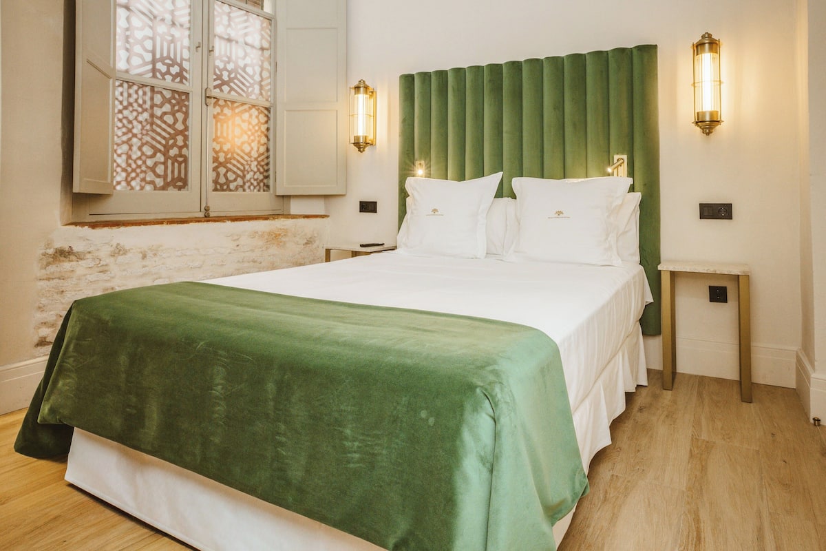 A neatly made bed with a green bedspread, a green headboard, and two wall-mounted light fixtures.