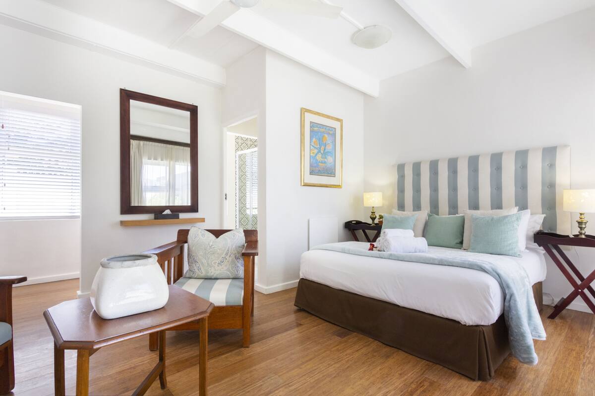 A well-lit bedroom with a large bed, a wooden chair, a small table, and a mirror on the wall.