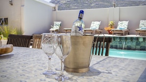 Uma mesa com uma garrafa de champanhe e duas taças, piscina e espreguiçadeiras com montanhas ao fundo.