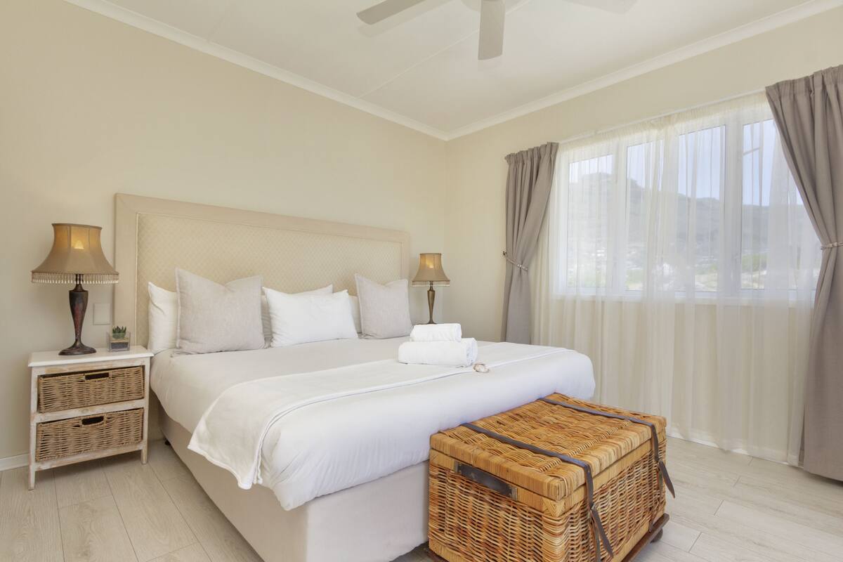 A hotel room with a large bed, two bedside tables, a nightstand, a wicker chest, and a view of the outdoors through sheer curtains.