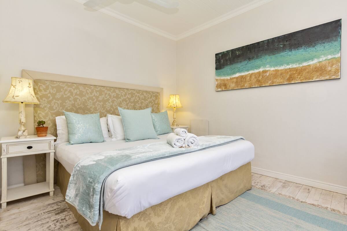 A neatly arranged bedroom with a bed, two bedside tables, lamps, a painting, and a small potted plant.