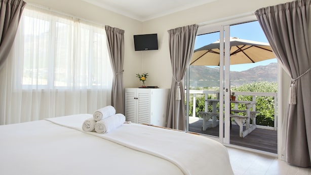 Quarto de hotel com uma cama grande, uma televisão e uma porta de vidro deslizante para um terraço ao ar livre com mesa e cadeiras.