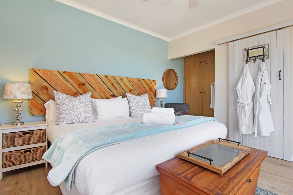 A bedroom with a wooden headboard, a bed with white bedding, a bedside table with a lamp, and a wooden chest of drawers.