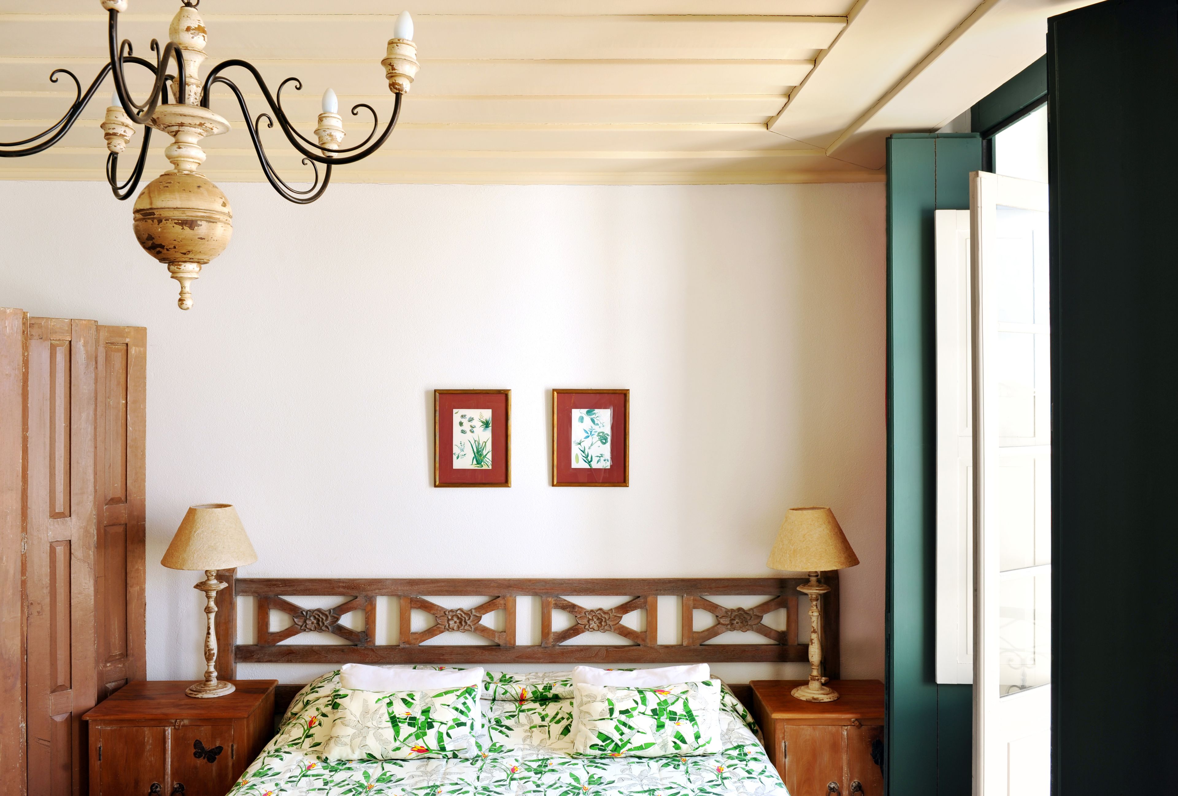 A bedroom with a wooden headboard, two bedside tables with lamps, a chandelier, and two framed botanical prints on the wall.