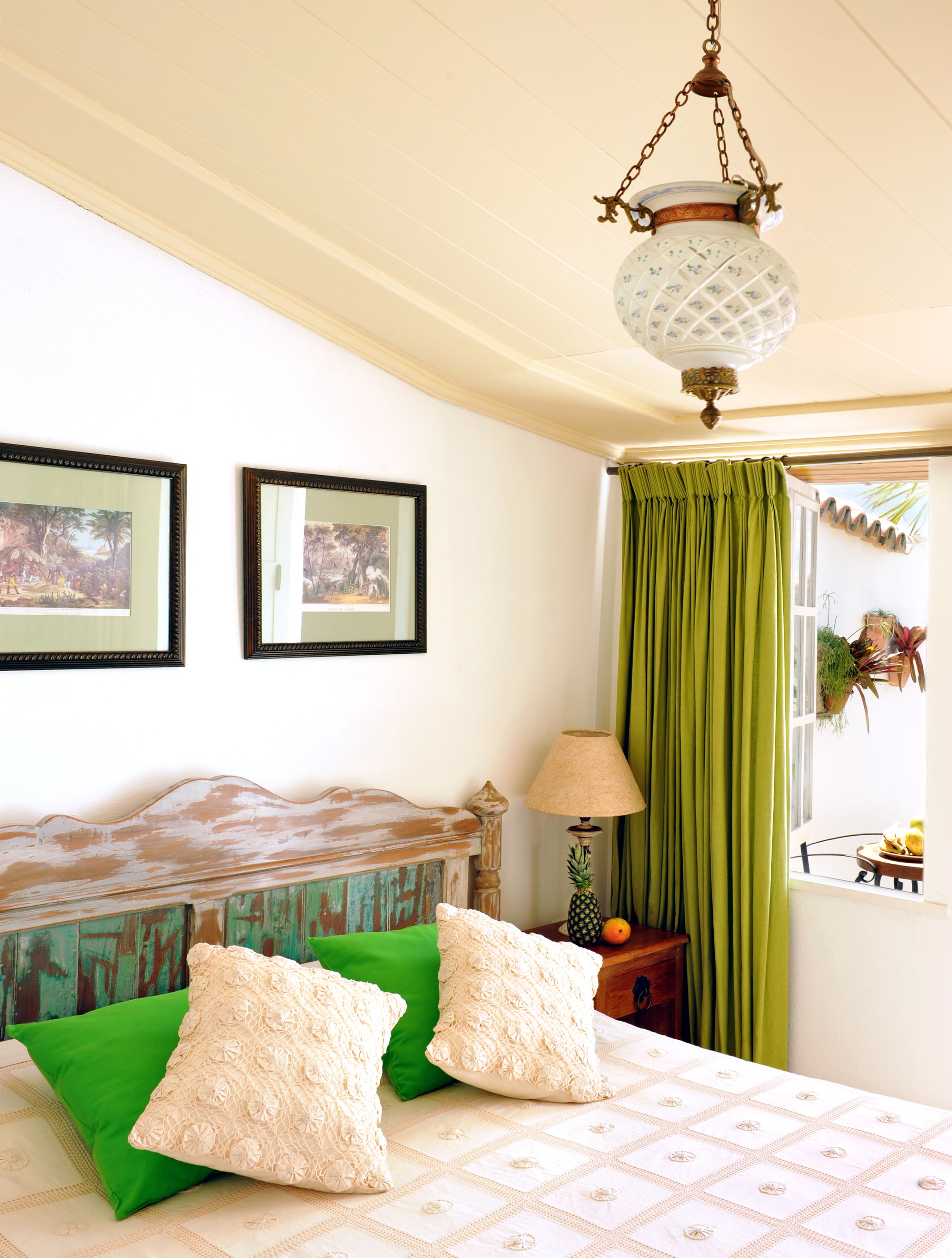 A bedroom with a bed, green pillows, a wooden headboard, a hanging lamp, a window with curtains, and framed pictures on the wall.