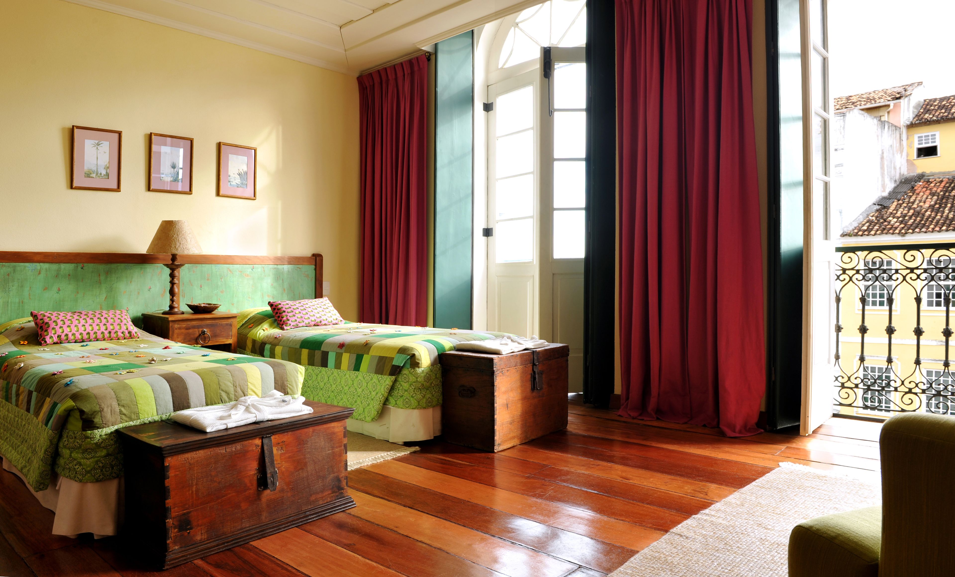 Quarto com duas camas, um baú de madeira, uma mesa de cabeceira e vista para uma varanda com grades de ferro forjado.