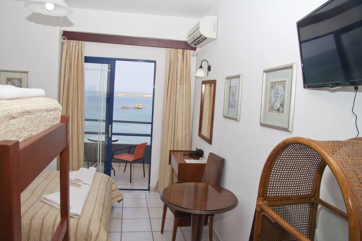 A hotel room with a bed, a dining table, a chair, a TV, and a view of the sea through a glass door.