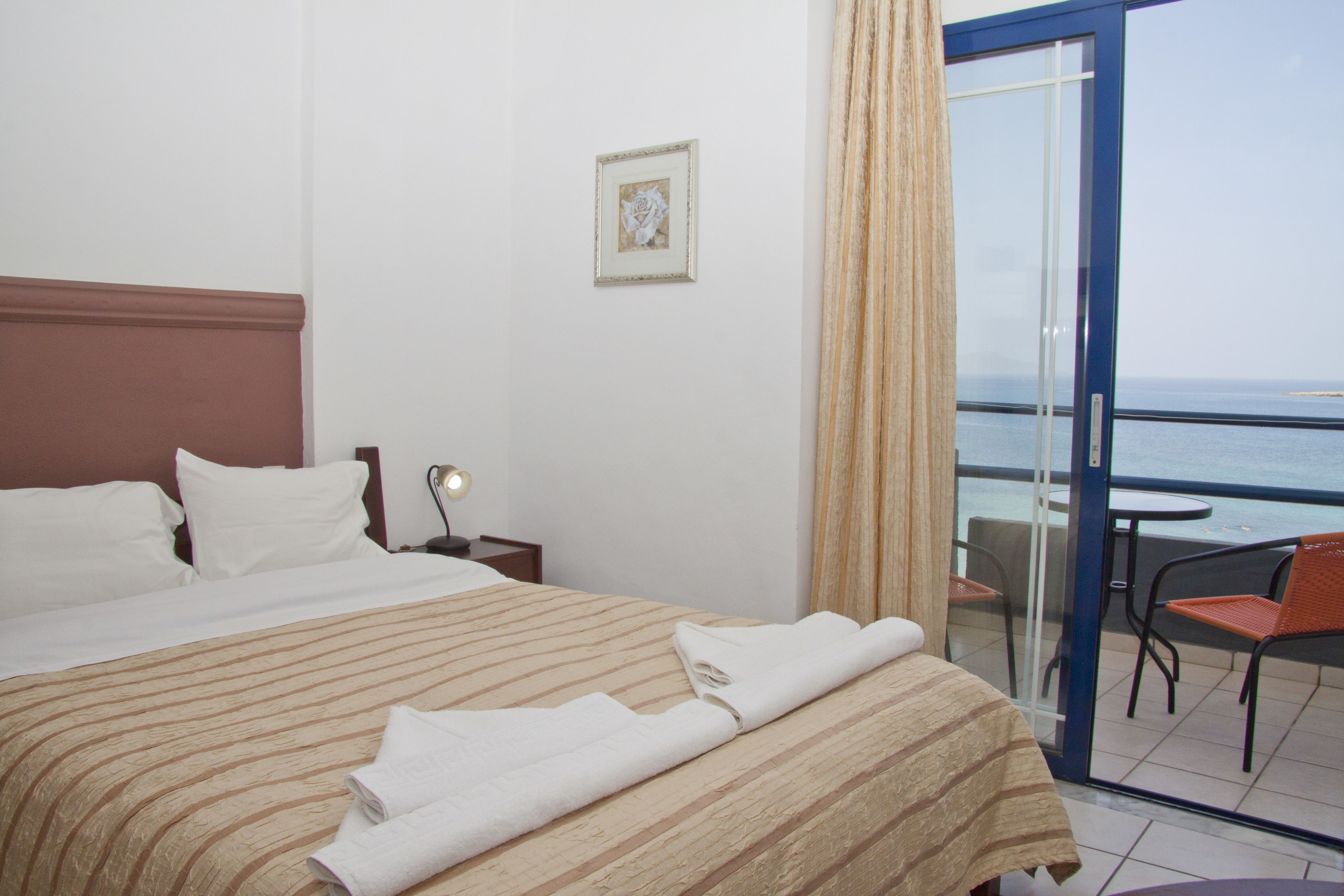 A hotel room with a bed, a nightstand, a framed picture on the wall, and a balcony with a table and chairs.