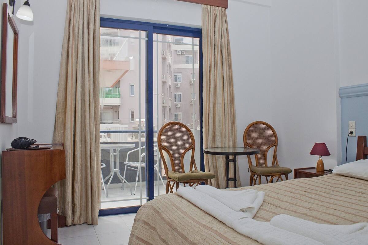 A hotel room with a bed, two wicker chairs, a small table, a lamp, and a view of buildings through a large window.