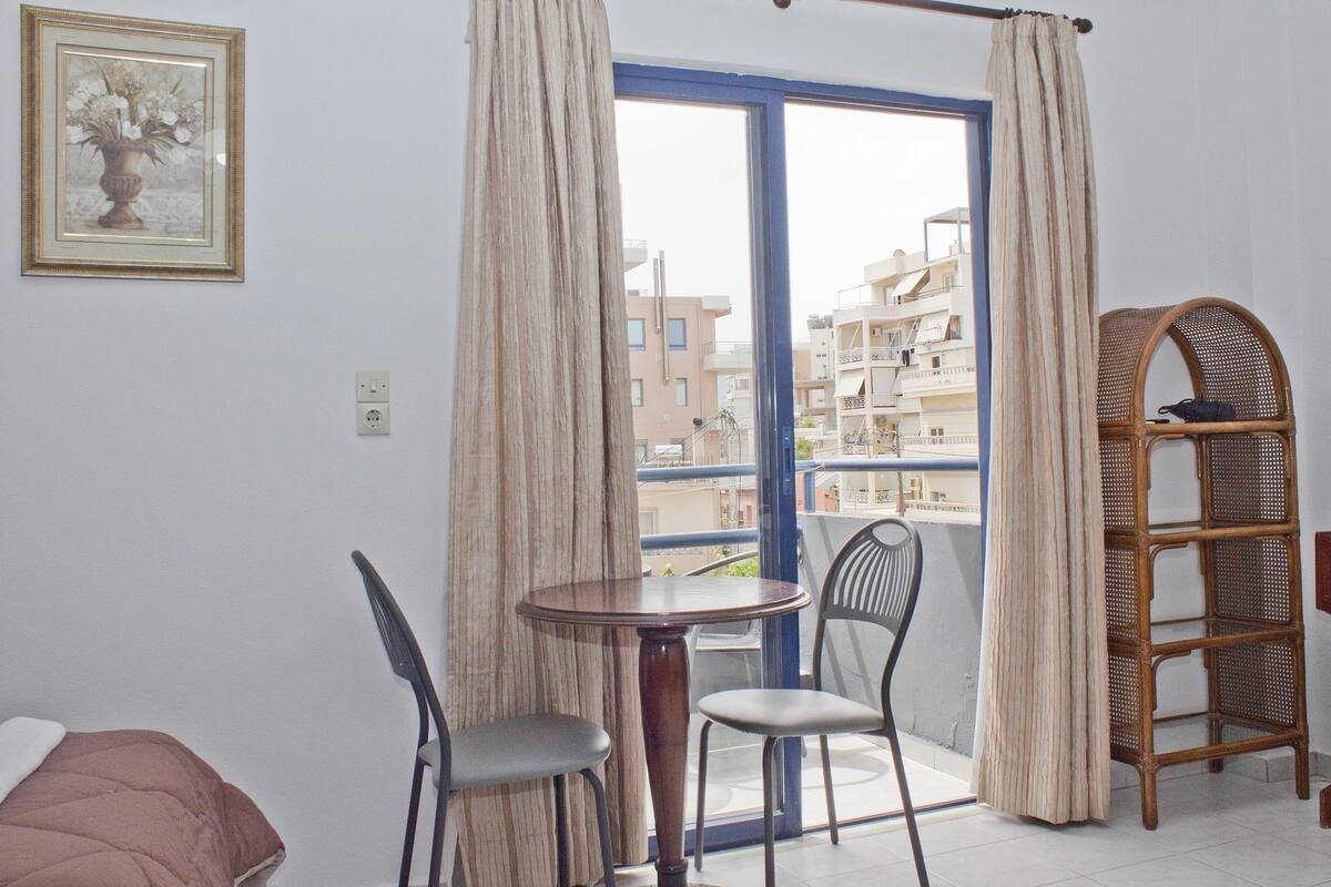 A room with a balcony, a dining table with chairs, a wicker chair, a bed, and a framed picture on the wall.