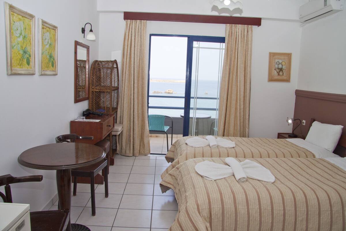 A hotel room with two beds, a desk, a chair, a small table, and a balcony with a view of the sea.