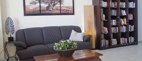 Una sala de estar con un sofá oscuro, una mesa de centro de madera con un florero, una estantería llena de libros y un cuadro de paisaje enmarcado en la pared.
