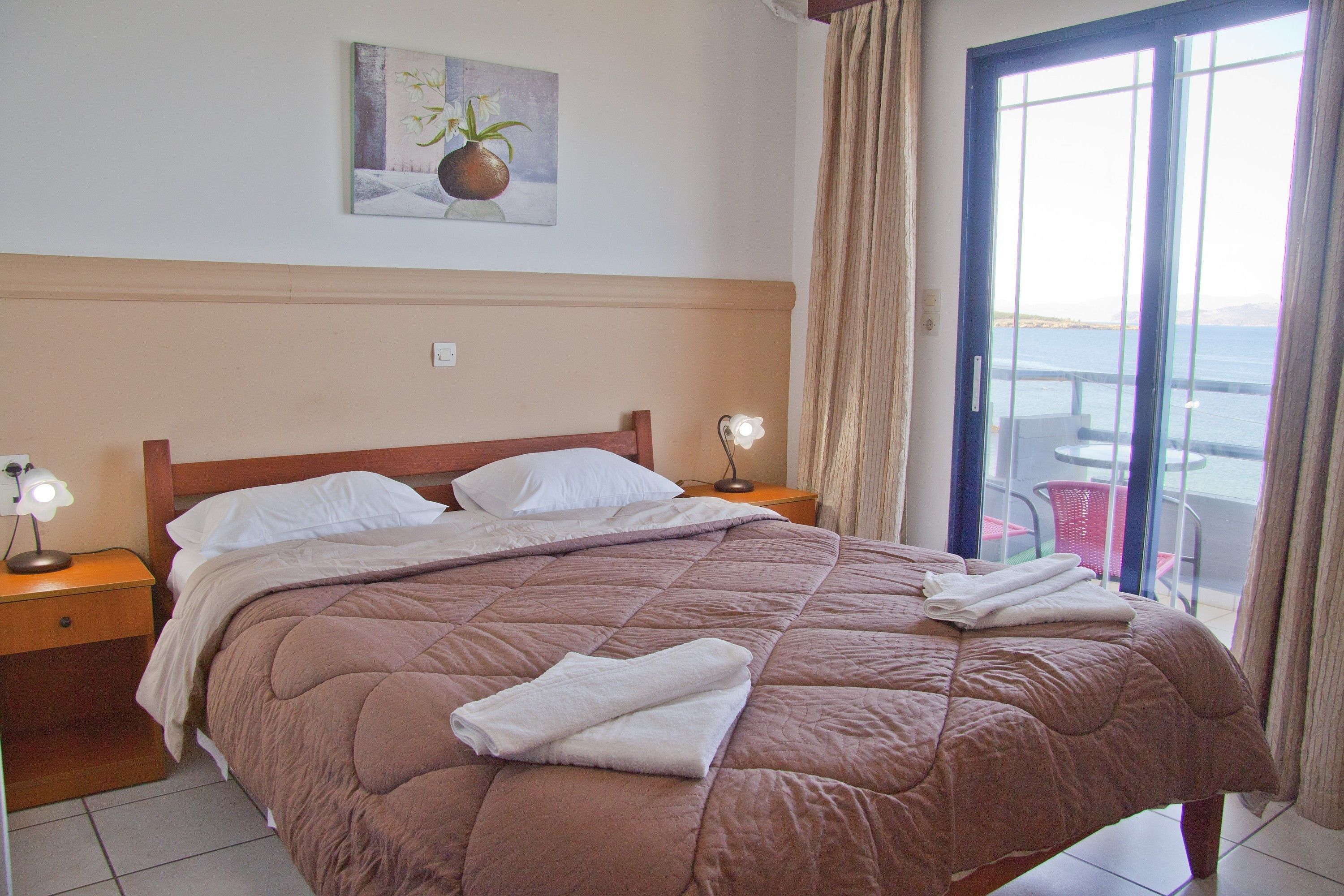A neatly made bed with a brown quilt, two white pillows, and folded towels on the side. There is a painting on the wall above the bed, and a window with curtains to the right.