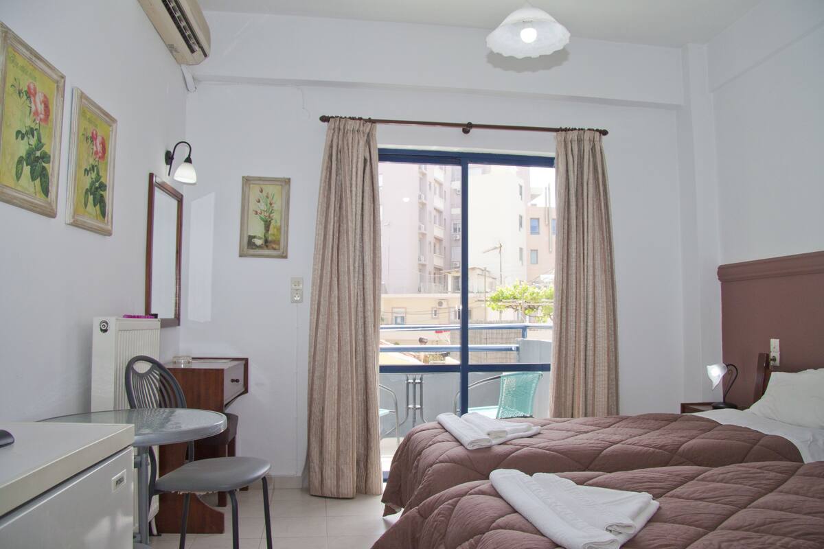 A hotel room with a bed, a desk with a chair, a window with curtains, and a view of buildings outside.