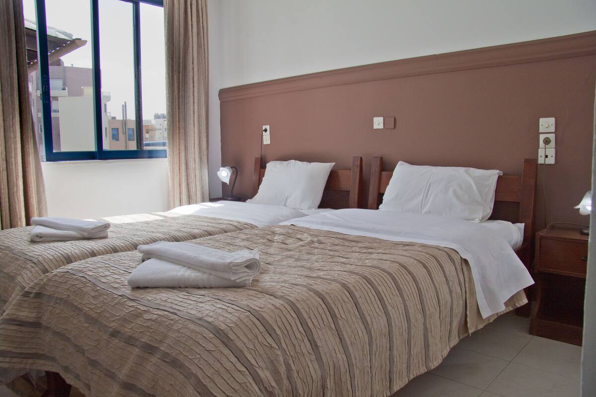 A hotel room with a large bed, two bedside tables, a window with curtains, and a view of buildings outside.
