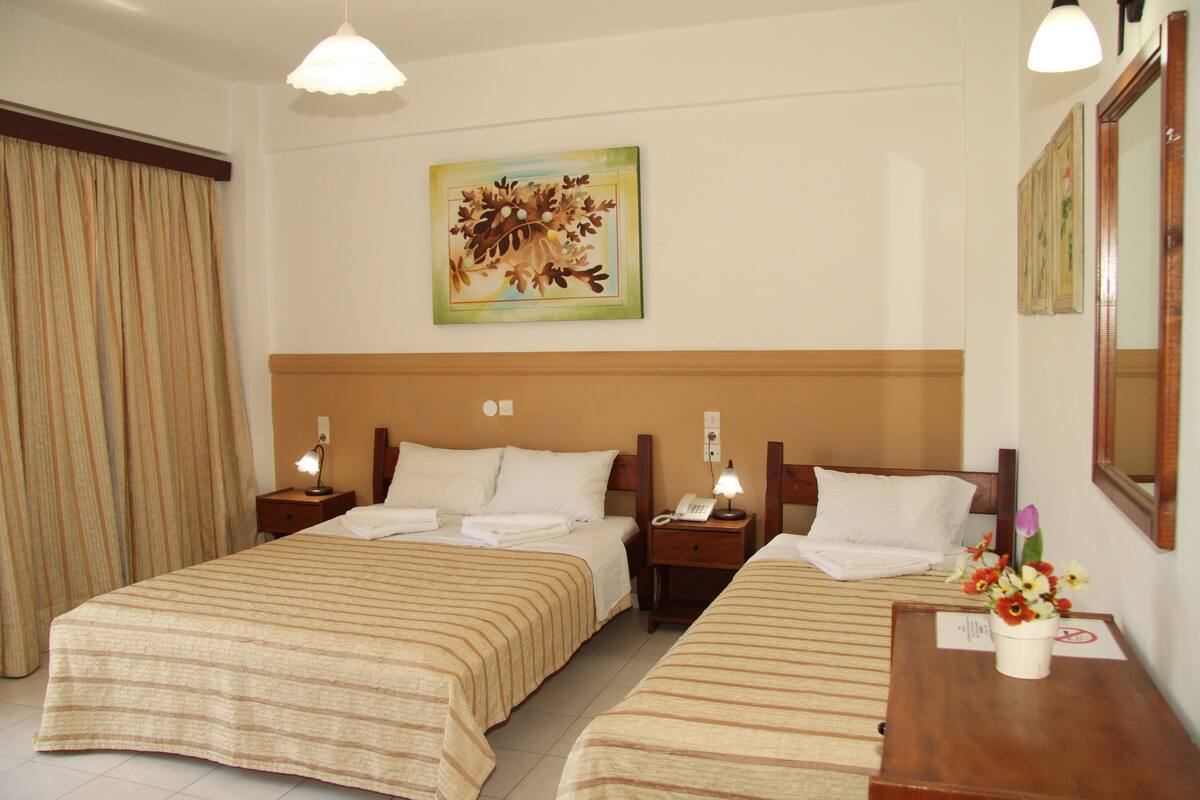 A hotel room with two beds, a painting on the wall, a nightstand with a lamp, and a vase of flowers on a table.