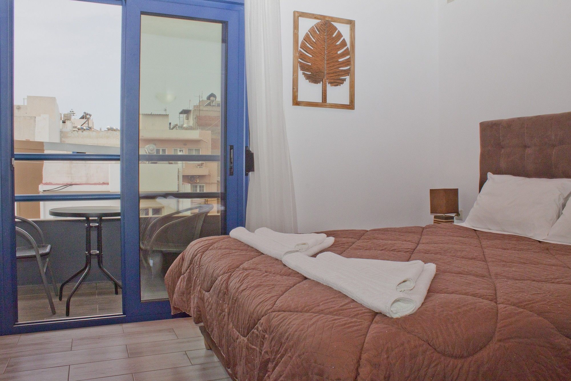 A hotel room with a bed, a small table with chairs, a balcony, and a leaf decoration on the wall.