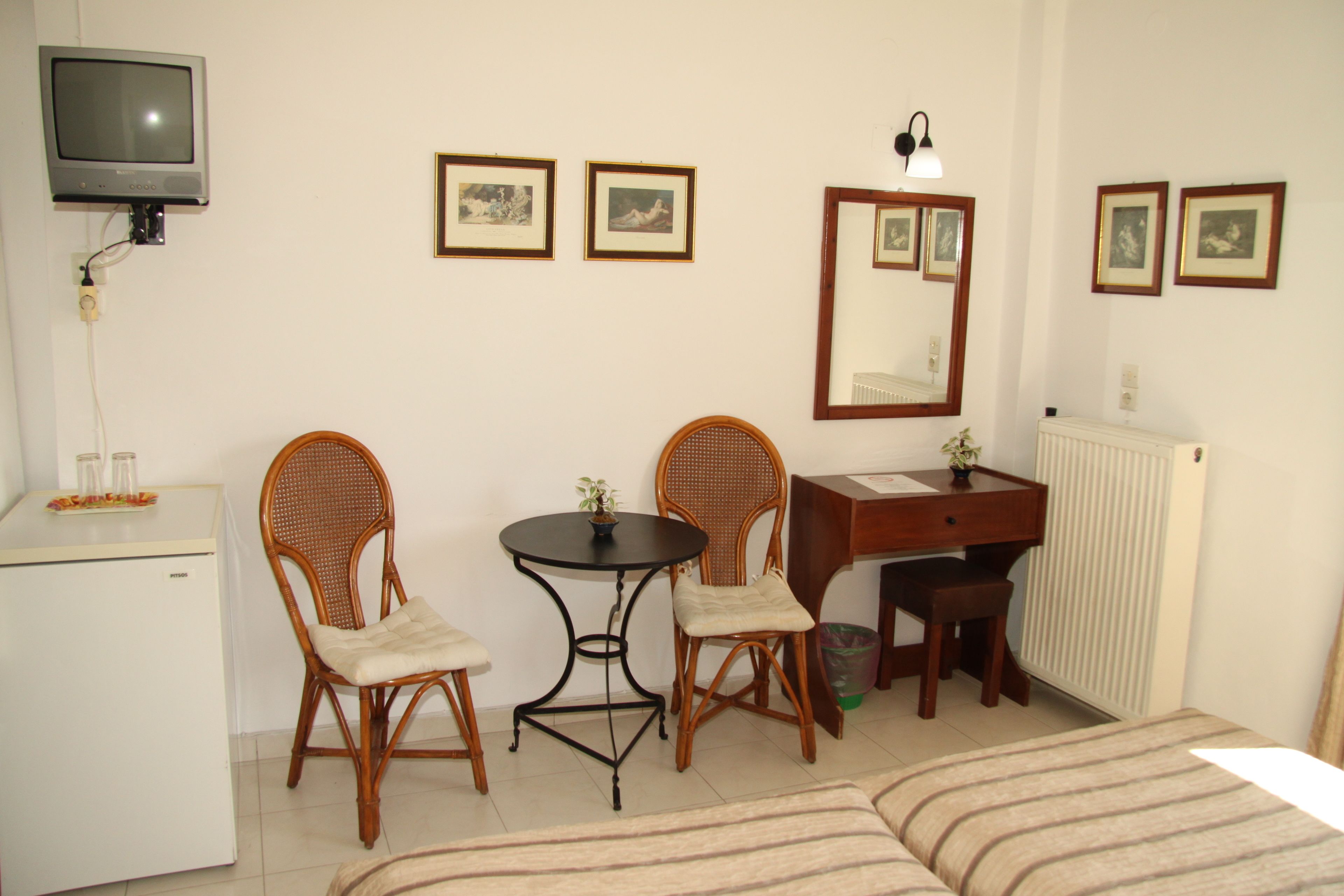 Una habitación pequeña y limpia con cama, dos sillones de mimbre, una mesita, escritorio, radiador y televisor en la pared.