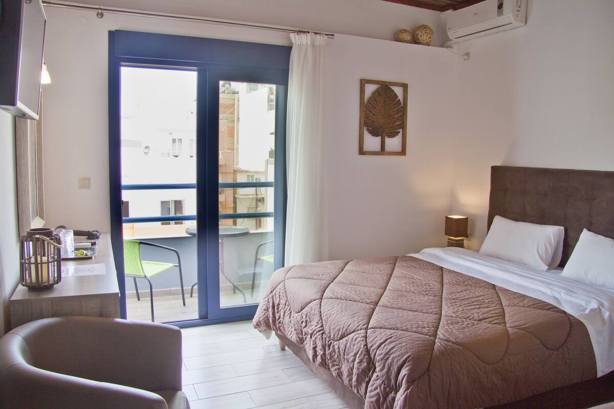 A hotel room with a bed, a chair, a desk with a lamp, and a view through a glass door.