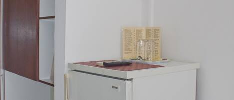 Un refrigerador blanco pequeño con la etiqueta “PITSOS” en un pasillo con paredes blancas y una puerta de madera.