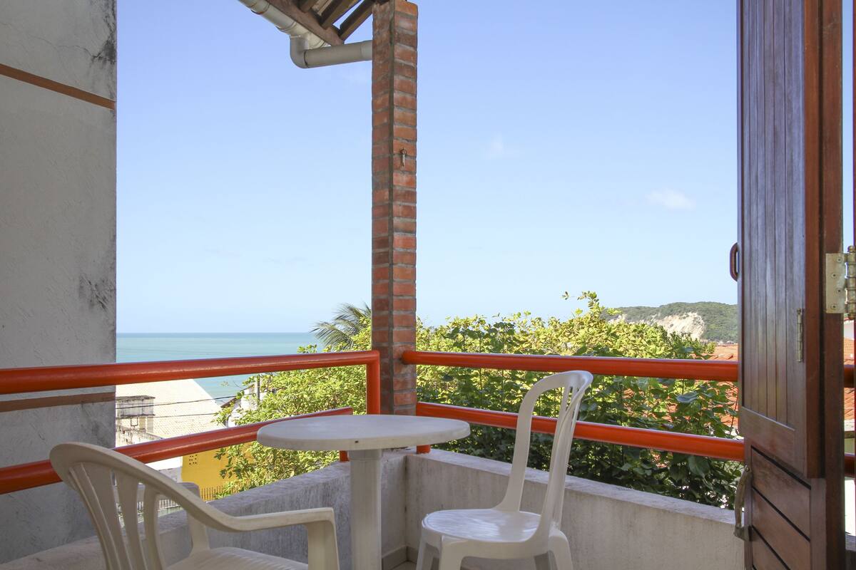 A balcony with a table and two chairs, overlooking a coastal view with greenery and hills.