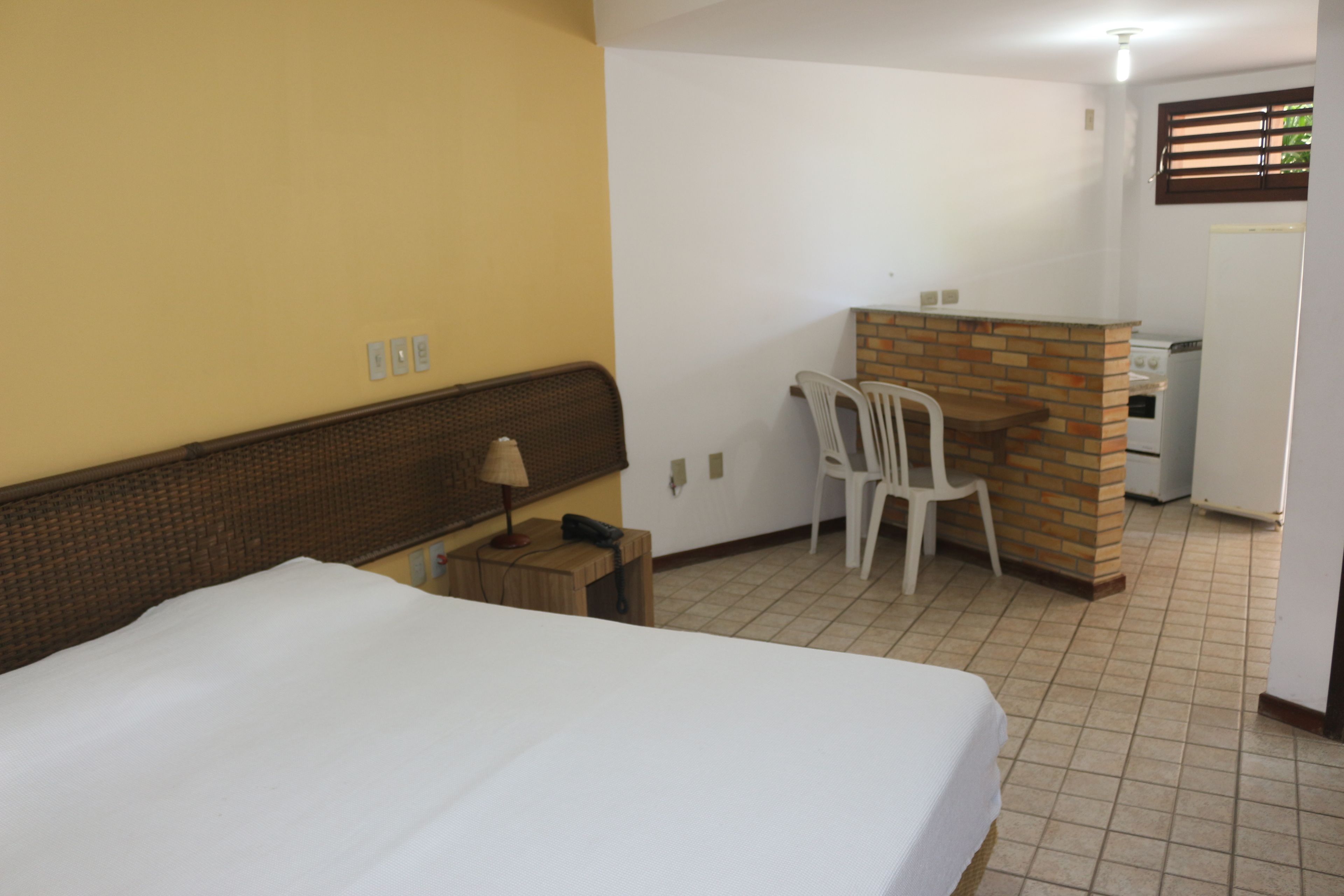 A hotel room with a bed, a small desk with a lamp, a chair, and a kitchenette area.