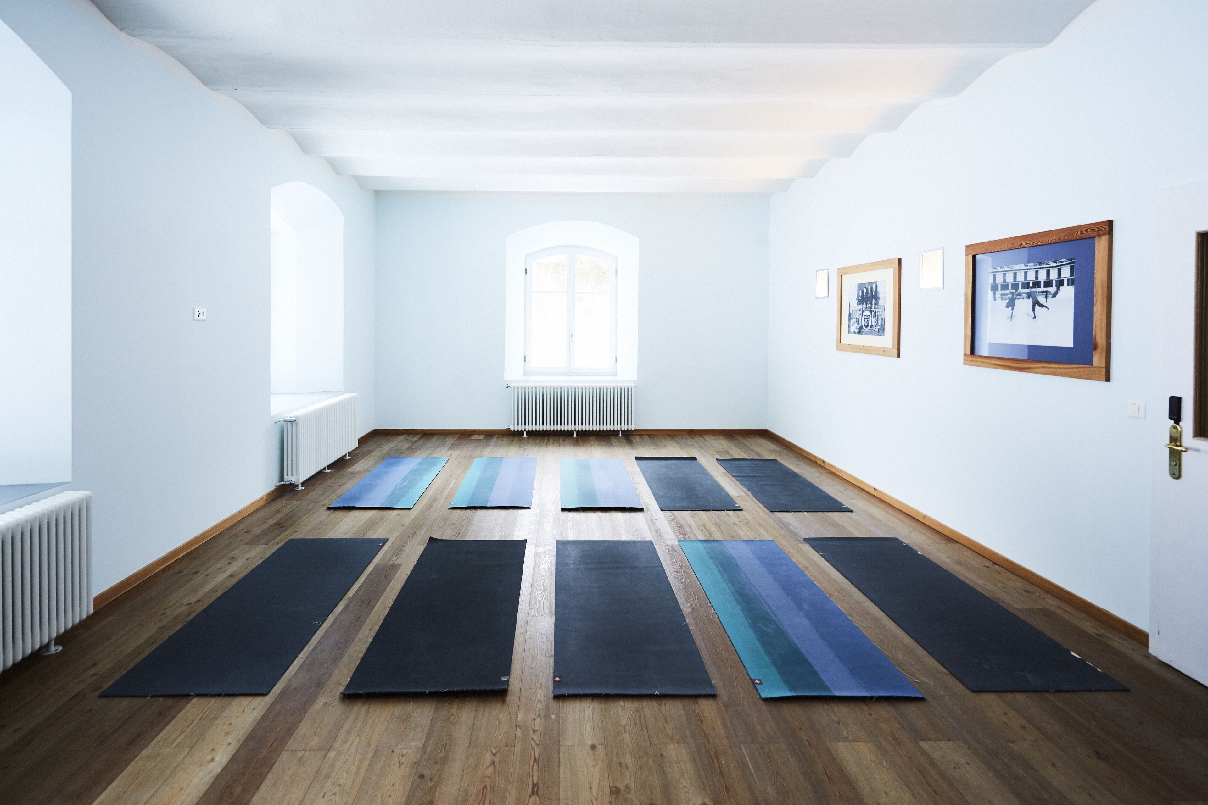 Un estudio de yoga con suelos de madera, esterillas de yoga negras y azules, un radiador y cuadros en la pared.