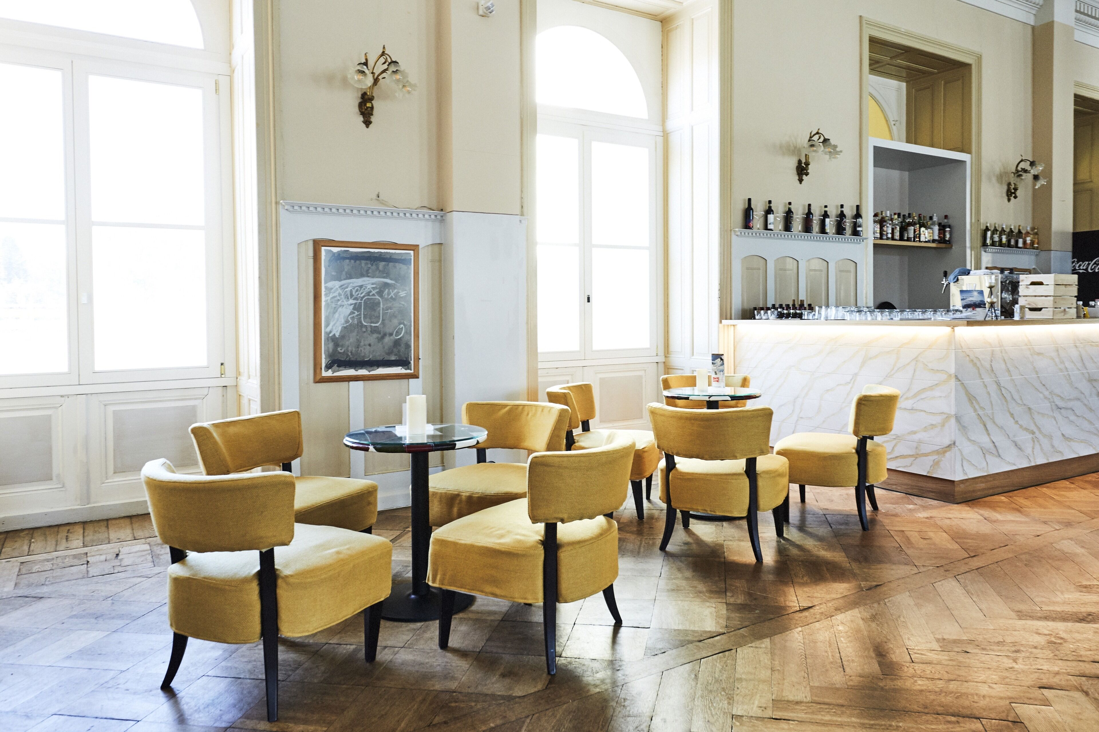 A spacious room with high ceilings, large windows, and a bar area with neatly arranged bottles.
