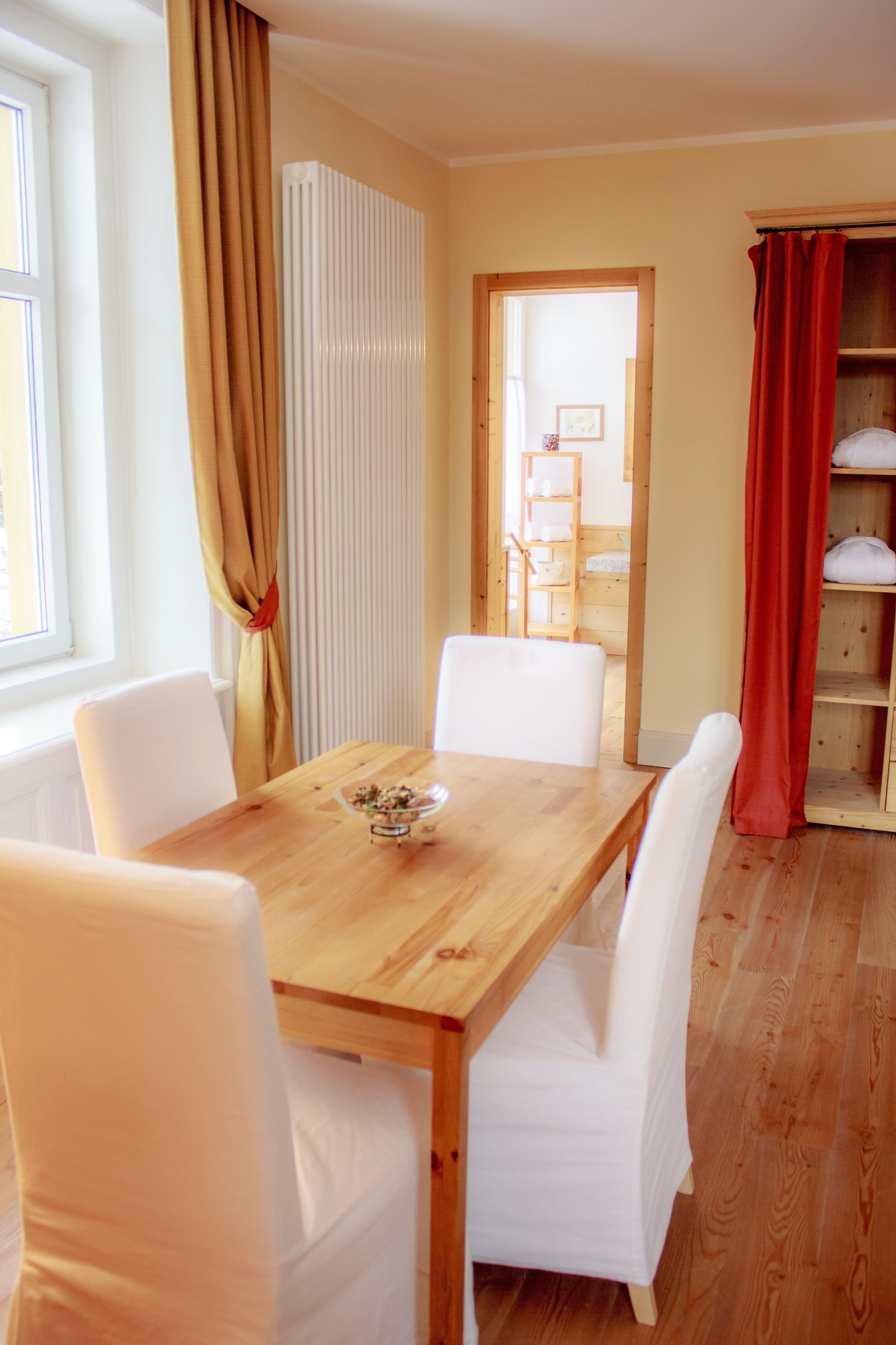 A dining area with a wooden table and white chairs, a radiator, and a red curtain.