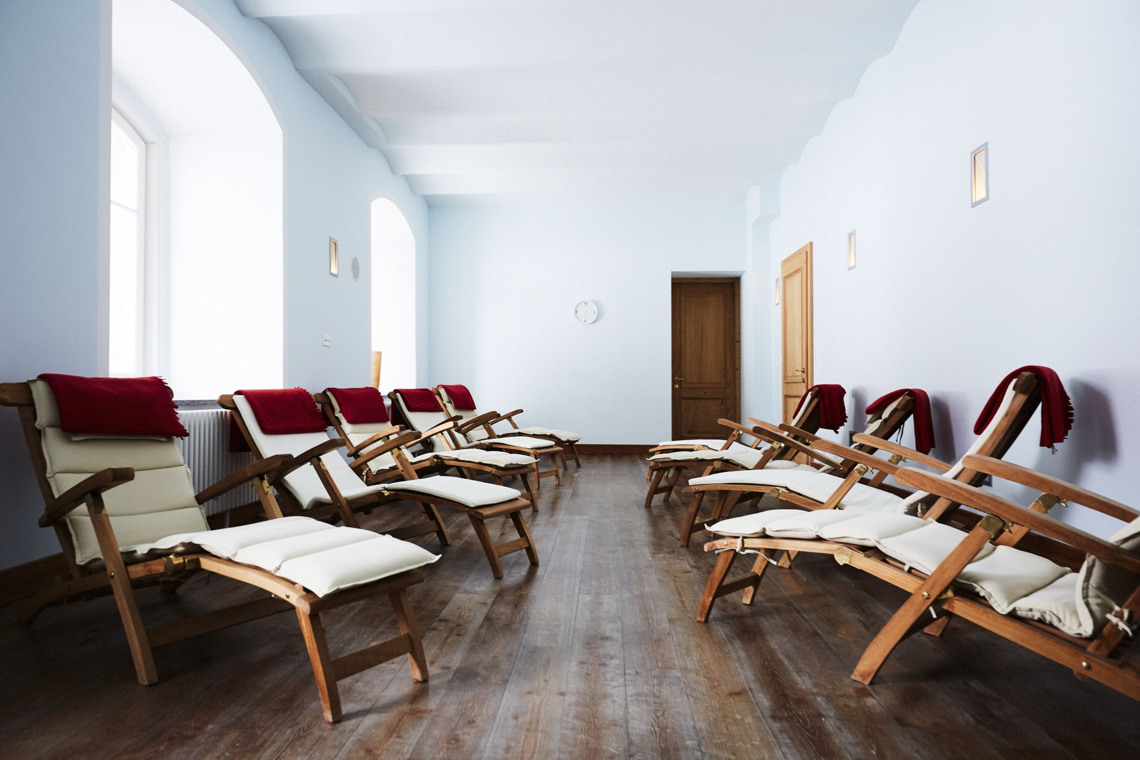 Een kamer met houten lounge fauteuils die in een halve cirkel zijn opgesteld, een houten deur en een radiator aan de muur.
