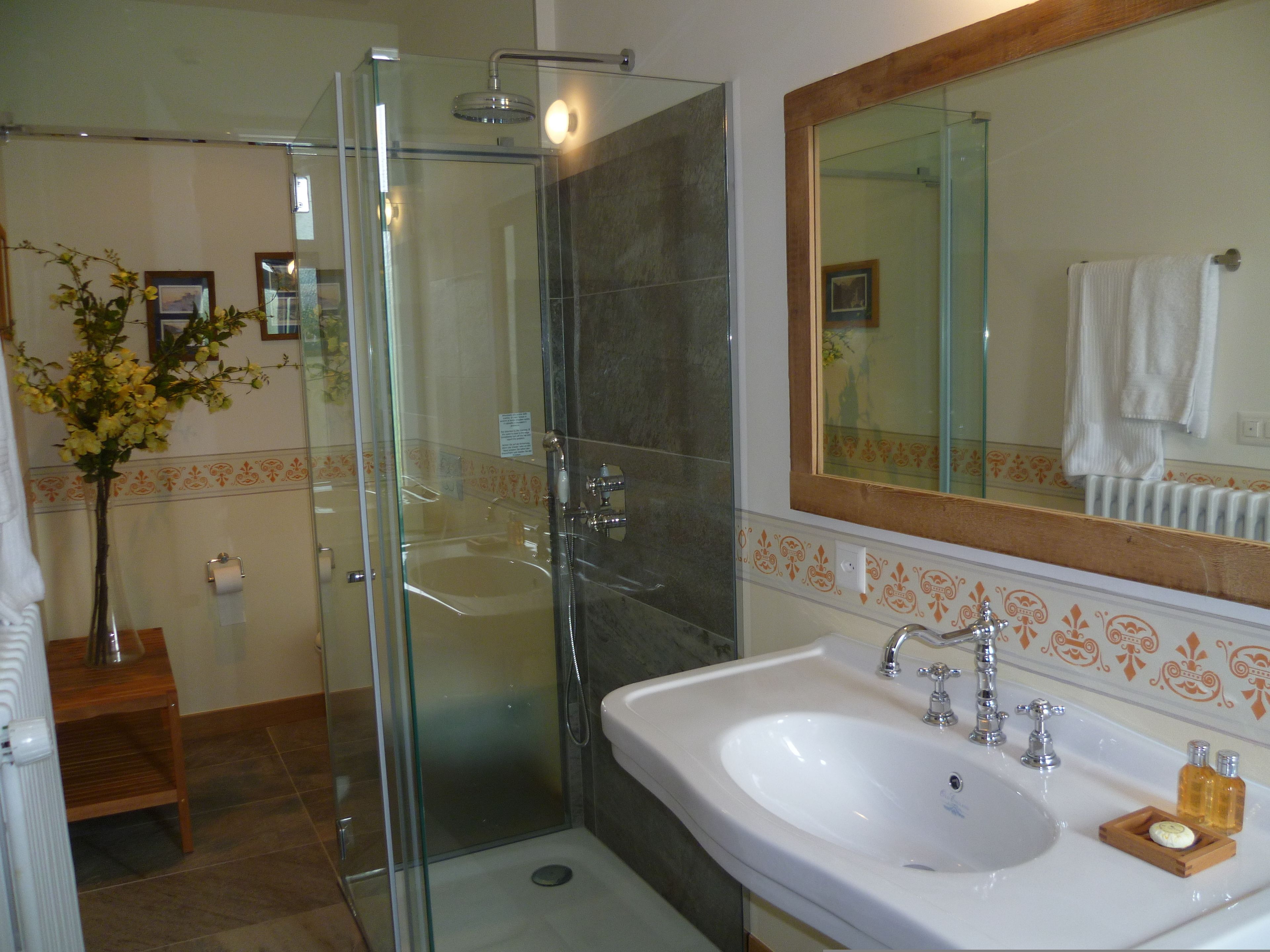 A bathroom with a glass shower enclosure, a white sink with a mirror above it, and a vase with flowers on a wooden stool.