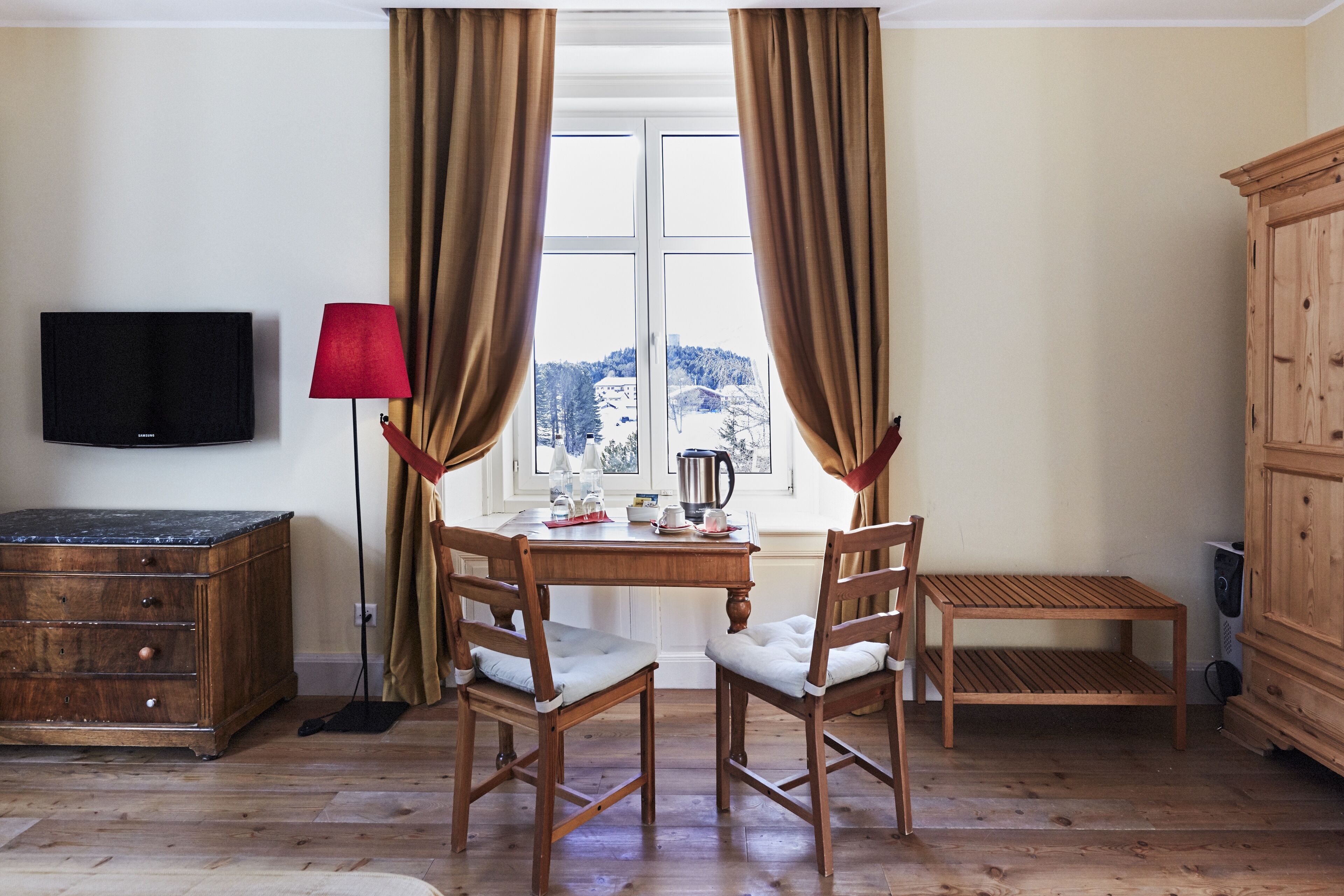 A room with a wooden table, two chairs, a television, a lamp, and a view of a snowy landscape through a window with curtains.