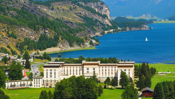 A large building with multiple floors and a prominent entrance, surrounded by lush greenery and a lake.